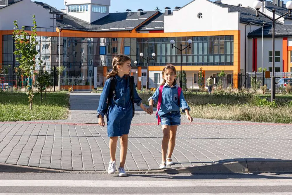 Enfants allant à l’école devant un bâtiment scolaire contemporain Deux écolières traversant la rue devant une école moderne avec sacs à dos
